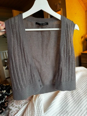 Stickad grå väst från Kaffe - Snygg stickad väst i grått från Kaffe. Västen är ärmlös, har ribbade detaljer längs sidorna och en öppen front. Perfekt att slänga över en skjorta eller t-shirt för en chill look. Materialet känns mjukt och lätt, vilket gör den enkel att bära hela dagen.