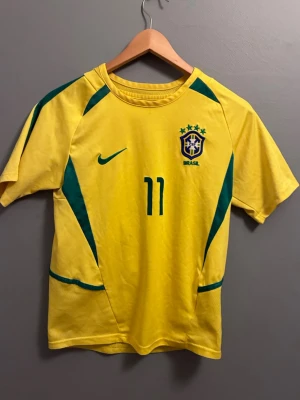 Brasilien fotbollströja Ronaldinho  - Gul Brasilien fotbollströja från Nike med gröna detaljer och nummer 11. På bröstet finns landslagets emblem och Nike-logga, på ryggen står det Ronaldinho och nummer 11. Perfekt för dig som älskar fotboll och vill stötta Brasilien. Är från fotbollsmarknaden