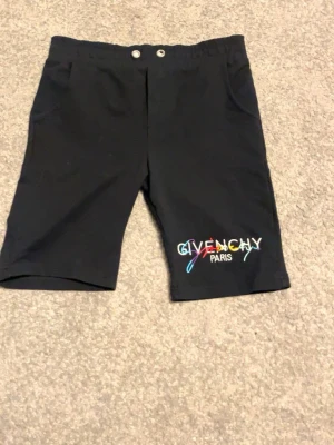Svarta shorts från Givenchy Paris - Snygga svarta shorts från Givenchy Paris med broderad logga i flera färger på ena benet. Klassisk passform med två knappar i midjan och fickor på sidorna. Perfekta för en trendig och avslappnad stil.