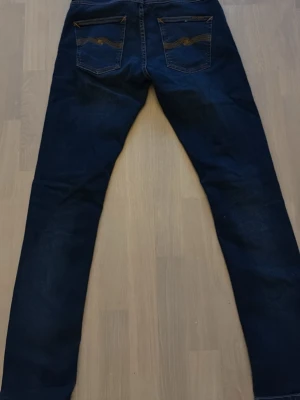 Mörkblå jeans från Nudie Jeans - Säljer ett par mörkblå jeans från Nudie Jeans med klassisk femficksmodell och kontrastsömmar på bakfickorna. Jeansen har rak passform och är tillverkade i slitstark bomull med en snygg tvättad look. Modellen är Grim Tim.
