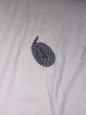 Svartvitt flätat tygbälte - Snyggt svartvitt flätat tygbälte med silverfärgat spänne och bruna läderdetaljer vid ändarna. Perfekt accessoar för att ge outfiten en cool touch. Bältet är flexibelt och passar till många olika stilar.