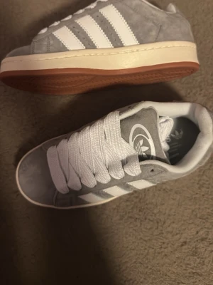 Grå adidas Campus sneakers i mocka - Snygga grå adidas Campus sneakers med klassiska vita ränder och vit sula. Ovandelen är i mjuk mocka och skorna har breda vita skosnören. Perfekta för dig som gillar en clean och stilren look.