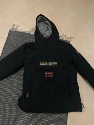 Svart vinterjacka Napapijri L - Fodrad, NY .Säljer en svart vindjacka från Napapijri med huva och stor ficka framtill. Jackan har dragkedjor på sidorna, norsk flagga på bröstet och Napapijri-logga ovanför. Perfekt för blåsiga dagar och snygg streetstil.