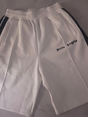 Vita shorts från Palm Angels - Säljer ett par vita shorts från Palm Angels med svart logga på vänster ben och svarta ränder längs sidorna. Shortsen har elastisk midja och sidofickor. Perfekta för en avslappnad och trendig look.