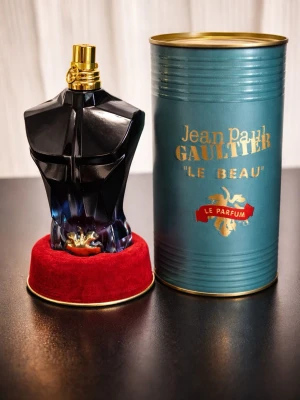 Jean Paul Gaultier Le Beau Le Parfum - Unik parfymflaska formad som en manlig torso i mörkblått glas med gulddetaljer och tillhörande turkos metalltub med guldfärgad text. Flaskan står på en röd sammetsliknande bas. En ikonisk doft från Jean Paul Gaultier.