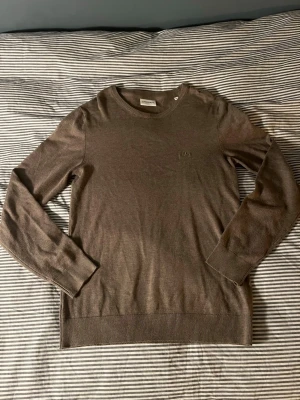 Brun stickad tröja från Lindbergh - Snygg brun stickad tröja från Lindbergh med rund halsringning och diskret broderad logga på bröstet. Tröjan har långa ärmar och ribbade muddar vid ärmslut och nederkant. Perfekt för lager på lager och enkel att matcha.
