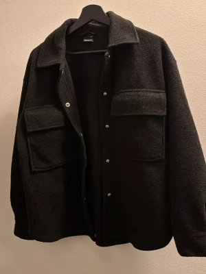 Svart overshirt/tröja från Gina Tricot - Säljer en svart overshirtjacka från Gina Tricot med stora bröstfickor och tryckknappar framtill. Jackan har en oversized passform och mjukt, fluffigt material.