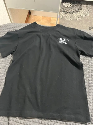 Svart Gallery Dept. t-shirt - Säljer en svart t-shirt från Gallery Dept. med vit logga tryckt på bröstet. Klassisk passform med rund hals och korta ärmar. Tillverkad i mjuk bomull som känns skön mot huden. Perfekt för dig som gillar streetwear och enkel stil.