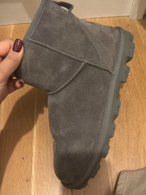 Gråa UGG tofflor i mocka - Mysiga gråa tofflor/skor från UGG i mjuk mocka med grov sula. Klassisk rund tå och diskret UGG-logga bak på hälen. Perfekta för att hålla fötterna varma under kyliga dagar. Använt max 3 ggr