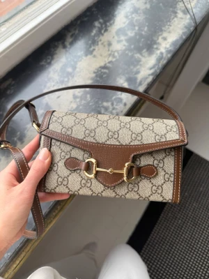 Gucci beige och brun axelväska - Säljer en snygg axelväska från Gucci i beige canvas med klassiskt GG-mönster och bruna skinn-detaljer. Väskan har guldfärgade metalldetaljer framtill och justerbar axelrem. Perfekt storlek för det viktigaste och stängs med magnetlås. Modell: Gucci horsebit 1955 mini bag. Nypris: 13 300kr