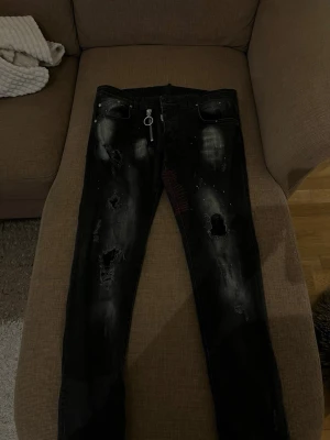 Svarta jeans med dragkedja och slitningar - Säljer ett par svarta Dsquared2 jeans med coola slitningar, färgstänk och en stor silverfärgad dragkedja framtill. Jeansen har en slim passform och detaljer som ger en edgy streetkänsla. Perfekta för dig som vill sticka ut med din stil.