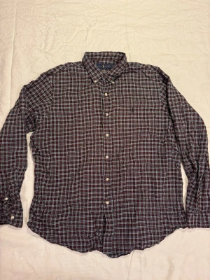 Rutig skjorta från Ralph Lauren XL - Klassisk rutig skjorta från Ralph Lauren i mörka toner av rött, blått och vitt. Skjortan har lång ärm, knappar framtill och en liten broderad logga på bröstet. Perfekt för dig som gillar tidlös stil och vill ha något som funkar till det mesta.