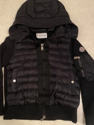 Svart dunjacka med huva från Moncler - Snygg svart dunjacka från Moncler med huva och dragkedja framtill. Jackan har två fickor med tryckknappar, ribbade muddar och en Moncler-logga på ärmen. Perfekt för kalla dagar och ger en sportig, trendig look.