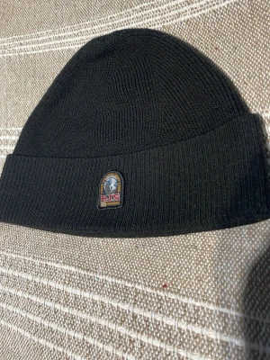 Svart ribbstickad parajumpers mössa - Svart ribbstickad mössa från parajumpers med uppvikt kant och en liten patch framtill med logga och text. Klassisk modell som sitter tajt på huvudet och passar perfekt till kyliga dagar. Materialet känns mjukt och stretchigt.