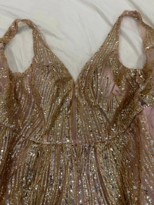 Glittrig långklänning i guld och rosa - Magisk långklänning med glittriga paljetter i guld och rosa toner. Klänningen har v-ringning, smala axelband och ett svepande fall. Perfekt för bal eller festliga tillfällen. Materialet känns lätt och skimrande, vilket ger en lyxig känsla. Den var min syrras balklänning super fin och glittrig! Hör av er om det är några frågor