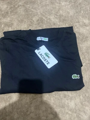 Svart t-shirt från Lacoste - Klassisk svart t-shirt från Lacoste med rund halsringning och den ikoniska gröna krokodilloggan broderad på bröstet. Tillverkad i mjuk bomull som känns skön mot huden. Perfekt för en stilren och enkel look. Den passar S/M