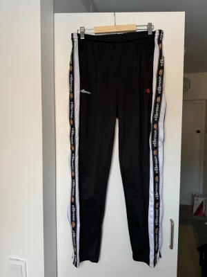 Svarta mjukisbyxor från Ellesse - Svarta mjukisbyxor från Ellesse med vita och svarta sidopaneler och logga längs benen. Byxorna har resår i midjan och är tillverkade i ett glansigt syntetmaterial. Perfekta för en sportig och avslappnad stil.