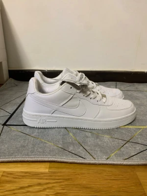 Nike Air Force 1 vita sneakers - Klassiska helvita Nike Air Force 1 sneakers med låg profil och rund tå. Skorna har snörning, perforerade detaljer på tån och är tillverkade i slätt skinn. Perfekta för dig som gillar en clean och tidlös stil.