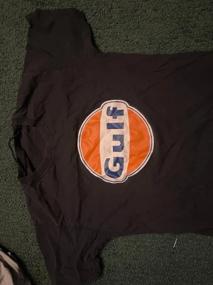 Svart Gulf t-shirt med tryck - Cool svart t-shirt med stort Gulf-tryck i orange, vitt och blått på bröstet. Klassisk rund hals och korta ärmar. Perfekt för dig som gillar retro och streetstyle.