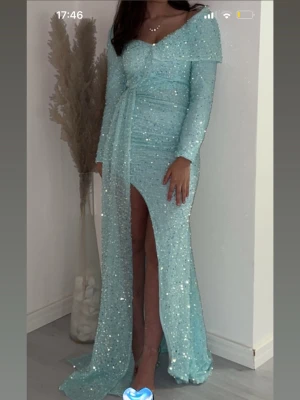 Ljusblå glittrig långklänning - Magisk ljusblå långklänning med glittriga paljetter, v-ringad urringning och lång ärm. Klänningen har en markerad midja med knytband och en hög slits framtill som ger en elegant look. Perfekt för bal eller festliga tillfällen.