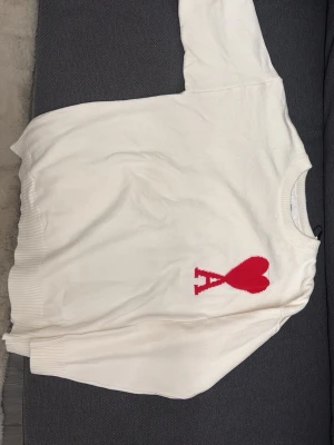 Vit stickad tröja från Ami Paris - Vit stickad tröja från Ami Paris med röd hjärtlogga och bokstaven 'A' på bröstet. Tröjan har rund halsringning, ribbade muddar vid ärmslut och nederkant samt en avslappnad passform. Perfekt för dig som gillar stilrena plagg med en twist.