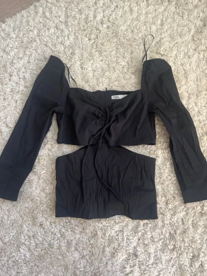 Svart topp från Zara - Svart topp från Zara med lång ärm och croppad passform. Har snörning framtill, en cut-out under bysten och dragkedja på baksidan. Använd typ 2 gånger💕 Säljer då den inte kommer till användning! Skriv om frågor💕