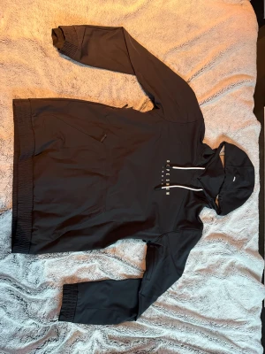 Svart vindjacka från Dope Snow - Svart vindjacka från Dope Snow med huva och vit snörning. Stor grafisk print på ryggen med berg i blått och rosa samt sol i vitt. Jackan har fleecefoder, elastiska muddar och praktisk magficka med dragkedja. Perfekt för dig som gillar street och outdoor.