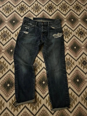 Mörkblå jeans Abercrombie & Fitch 32/34 - Snygga mörkblå jeans från Abercrombie & Fitch med slitna detaljer och råa kanter vid benslut. Klassisk femficksmodell med raka ben och diskreta slitningar på fram- och bakfickor. Perfekt för dig som gillar en avslappnad streetstil.