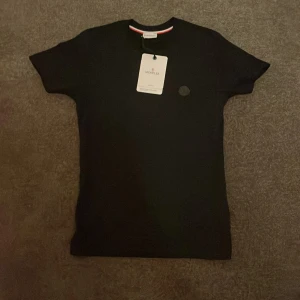 Svart Moncler t-shirt - Svart t-shirt från moncler. T-shirten är tillverkad i mjuk bomull och har korta ärmar. Snygg och stilren modell med en liten detalj i form av röd och vit rand på insidan av kragen.