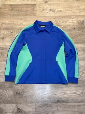 J.Lindeberg  - Snygg overshirt från J.Lindeberg i stark blå färg med mintgröna paneler längs sidor och ärmar. Skjortan har klassisk krage, tryckknappar framtill och diskret logga på bröstet. Materialet är mjukt och stretchigt polyester, perfekt för en sportig och modern look.