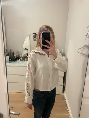 Vit feminin skjorta med spetsdetaljer  - Skjortan/blusen köpte jag second hand på stadsmissionen men den är från SHEIN, inga tydliga tecken på användning. Den har långa ärmar och snygga spetsdetaljer längs axlar och ärmar. Skjortan har klassisk krage, knäppning framtill och en lite loose passform. Perfekt för dig som gillar en feminin och trendig look.
