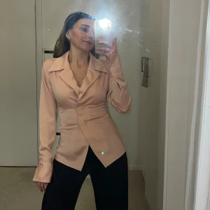 Rosa satinblus med v-ringning - Snygg satinblus från Gina Tricot med markerad midja, v-ringning och bred krage. Lax-rosa färgad. Långa ärmar med knappar vid manschetten och två dekorativa knappar framtill. Perfekt för dig som gillar en stilren och trendig look. Helt ny oanvänd, endast tvättad. 