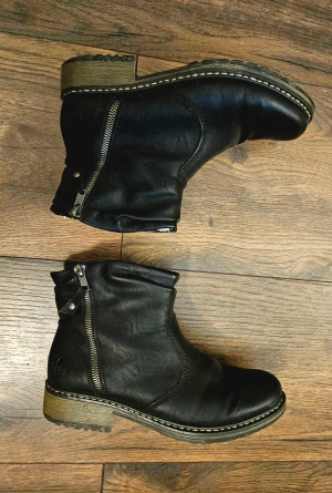 Svarta boots från Rieker med dragkedja - Svarta boots från Rieker med rund tå och grov platt sula. Utsidan är i skinn och insidan är fodrad med mjuk, vit syntetpäls för extra värme. Dragkedja på sidan gör dem enkla att ta på. Snygga kontrastsömmar runt sulan och robust look.