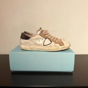 Philippe Model sneakers i beige och vitt - Snygga sneakers från Philippe Model i beige och vitt med svarta detaljer. Skorna har en klassisk låg siluett, snörning och en platt sula. Ovandelen är i en mix av skinn och mocka, med kontrasterande paneler och en mörk hälkappa. Perfekta för en avslappnad och trendig look. Skicket är som på bild finns mycket kvar i dem och har lite defekter, defekterna är som på bild box och tags medföljs 