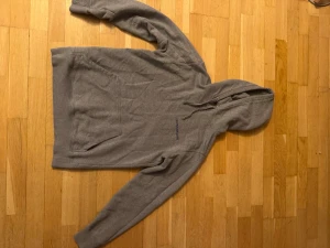 Grå hoodie från Calvin Klein Jeans - Säljer en grå hoodie från Calvin Klein Jeans i storlek M. Tröjan har huva med snörning, magficka och blått tryck på bröstet. Mjuk och skön bomullsmix, perfekt för chill dagar. Klassisk passform och långa ärmar.