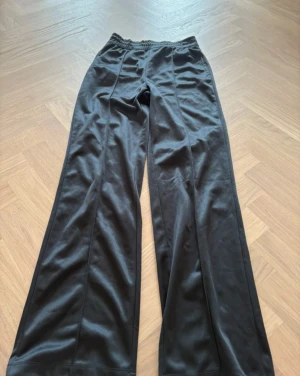 Svarta wide mjukisbyxor med resår - Svarta mjukisbyxor med breda ben och elastisk midja. Materialet har en lätt glansig finish och känns mjukt mot huden. Snyggt sömmade längs benen för extra stilpoäng. Perfekt för chill dagar eller när du vill ha en relaxed vibe.