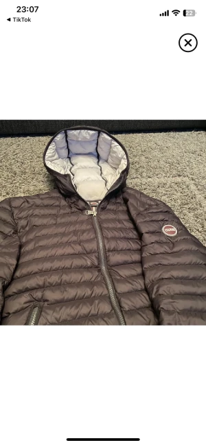 Säljer nu min snygga Colmar jacka  - Snygg svart pufferjacka från Colmar med huva och ljusgrått foder. Jackan har Colmar-logga på ärmen, två fickor med dragkedja och quiltad design. Perfekt för dig som vill ha en stilren och varm jacka med cool streetkänsla.