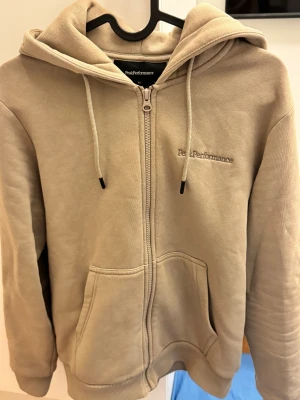 Peak Performance zip hoodie  - Säljer en beige hoodie från Peak Performance i storlek XS. Tröjan har dragkedja, huva med snörning och två stora fickor framtill. Insidan är mjuk och fleecefodrad för extra komfort. Perfekt för chill dagar och har diskret logga broderad på bröstet. Pris kan justeras något nypris 1500