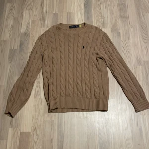 Beige kabelstickad tröja från Polo Ralph Lauren - Säljer en stilren beige kabelstickad tröja från Polo Ralph Lauren. Tröjan har en klassisk design med långa ärmar och ribbstickade muddar. Perfekt för en elegant och avslappnad look.