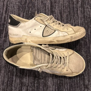 Philippe Model  - Säljer ett par vita sneakers från Philippe Model med grå detaljer och logga på sidan. Skorna har snörning med märkta skosnören och svart detalj vid hälen. Tillverkade i skinn med platt sula och rund tå, perfekta för en avslappnad och trendig look.