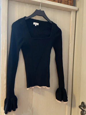 Svart ribbad topp från Stockh LM Studio - Svart långärmad topp från Stockh LM Studio i ribbad material. Fyrkantig halsringning och volangdetaljer vid ärmslut och nederkant i ljusbeige. Slim passform som sitter snyggt och ger en cool siluett. Perfekt för dig som gillar stilrena detaljer.