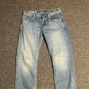 G-Star RAW MOSA Straight ljusblå jeans - Säljer ett par G-Star RAW MOSA Straight jeans i ljusblå tvätt med klassisk femficksdesign och raka ben. Jeansen har snygga slitningar framtill och tydliga sömmar, samt en RAW-logga på bakfickan. Perfekt för dig som gillar en clean och avslappnad look.