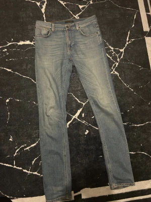 Ljusblå slim fit jeans från Nudie Jeans - Säljer ett par ljusblå slim fit jeans från Nudie Jeans med klassisk femficksdesign och snygga detaljer på bakfickorna. Jeansen har orangea sömmar och en mörkbrun patch med Nudie-loggan bak i midjan. Perfekt passform för dig som gillar smalare jeans. 34 längd 32 bredd 