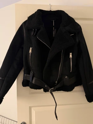 Svart teddyjacka med bälte och dragkedjor - Säljer en svart teddyjacka från Myriam-marcela med feta detaljer som bred krage, synliga dragkedjor och bälte i midjan.