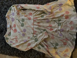 Klänning från Lindex med körsbärs print - Söt klänning i sobra färger från Lindex med rund hals och långa ärmar. Klänningen är ljusbeige med ett färgglatt körsbärs mönster i rosa, lila och grönt. Tillverkad i mjuk bomull med ribbad struktur, perfekt för vår och sommar men även vintertid med leggings.