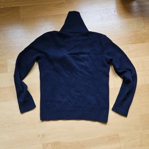 Mörkblå stickad tröja Polo Ralph Lauren - Mörkblå stickad tröja från Polo Ralph Lauren med hög krage och knappar vid halsen. Tröjan har lång ärm, ribbade muddar och en liten bröstficka med knapp. Perfekt för kyliga dagar och snygg till jeans.