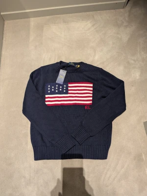 Mörkblå stickad tröja Polo Ralph Lauren - Snygg mörkblå stickad tröja från Polo Ralph Lauren med amerikansk flagga i vitt, rött och blått på bröstet samt RL-broderi. Tröjan har rund hals och långa ärmar med ribbade muddar. Perfekt för dig som gillar klassisk och cool stil. Stl M, för kille mer som S.