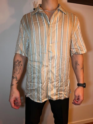 Beige och vit randig skjorta - Snygg kortärmad skjorta i beige med vita och blå ränder. Skjortan har klassisk krage, knäppning framtill och en loose fit som ger en avslappnad vibe. Perfekt för dig som gillar stilrena och fräscha plagg med lite retrofeeling. Stryckning 10kr 😂