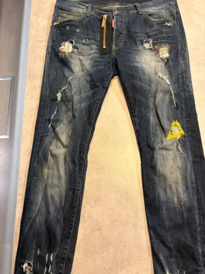 Dsquared2 blå jeans med dragkedja - Snygga blå jeans från Dsquared2 med slitna detaljer, färgstänk och en unik gul tryckt patch på ena benet. De har en cool dragkedja framtill och klassiska fem fickor. Passformen är rak och materialet är robust denim med kontrastsömmar.