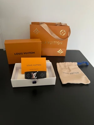 Louis Vuitton svart armband Monogram - Snyggt svart armband från Louis Vuitton med klassiskt monogrammönster och silverfärgad LV-logga. Tillverkat i exklusivt skinn med detaljer i silver. Kommer med original ask, dustbag och påse. Perfekt accessoar för dig som gillar lyx och stil.